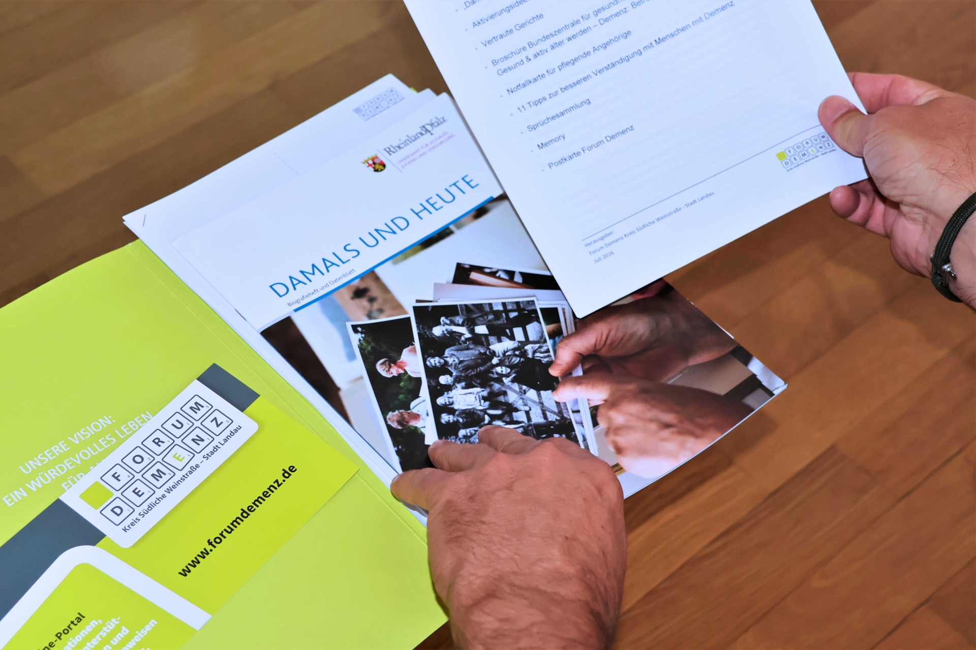 Mehrere Broschüren und Informationsunterlagen zum Thema Demenz liegen auf einem Holztisch. Im Mittelpunkt befindet sich die Broschüre "Damals und heute". Eine Person hält ein Informationsblatt in der Hand und blättert durch die Unterlagen.