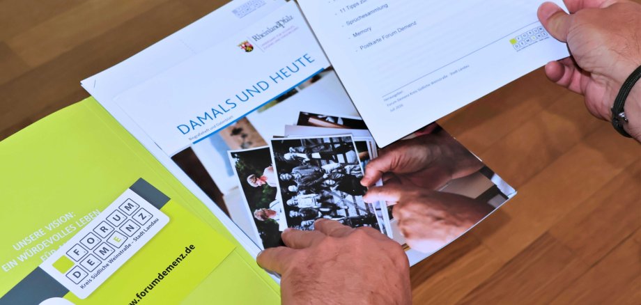 Die Aktivierungsmappe enthält unter anderem ein Biografieheft, einfache Koch- und Backrezepte, ein Memoryspiel und zahlreiche Beschäftigungsideen für den Alltag. Außerdem finden sich darin eine Notfallkarte für pflegende Angehörige sowie Informationen zu Beratungs- und Unterstützungsangeboten für Menschen mit Demenz und ihre Familien. Foto: Stadt Landau Mehrere Broschüren und Informationsunterlagen zum Thema Demenz liegen auf einem Holztisch. Im Mittelpunkt befindet sich die Broschüre "Damals und heute". Eine Person hält ein Informationsblatt in der Hand und blättert durch die Unterlagen.