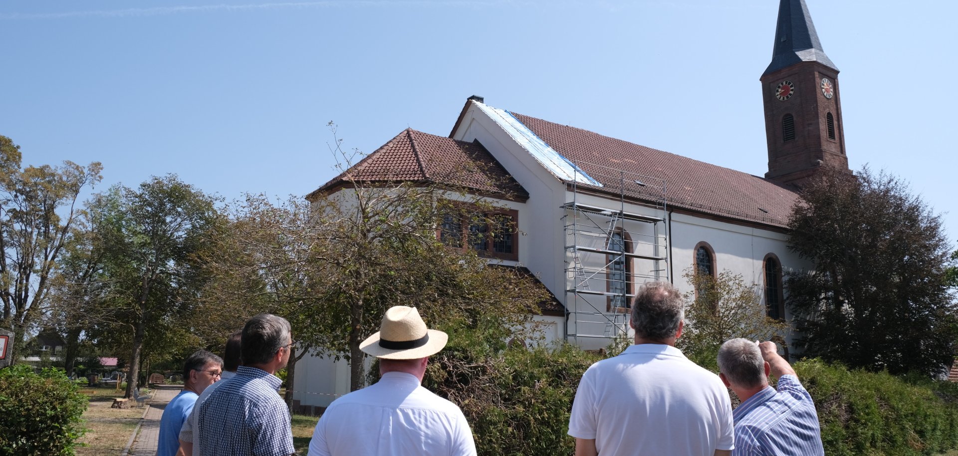 Eine Gruppe von Menschen betrachtet bei sonnigem Wetter eine Kirche von außen. Das Gebäude mit hellem Kirchenschiff, roten Ziegeln und hohem Kirchturm ist teilweise eingerüstet.