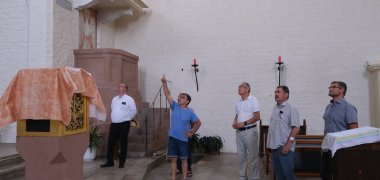 Pfarrer Stefan Häußler (Pfarrei Hl. Edith Stein Bad Bergzabern mit Kapsweyer, links), Stefan Zirker (Vorsitzender des Gemeindeausschusses der Kirche, hinten verdeckt), Markus Glaser (Verwaltungsrat der Pfarrei Edith Stein, zuständig für Kapsweyer, blaues T-Shirt), Ortsbürgermeister Bernhard Jud (2. von rechts) und Heiko Angemeier (Erster Beigeordneter der Ortsgemeinde), dem Landrat Dietmar Seefeldt (3. von rechts), welche Schäden das Unwetter im Inneren des Gotteshauses verursacht hat. Foto: KV SÜW Mehrere Männer stehen im Innenraum einer Kirche und betrachten Schäden am Dach.