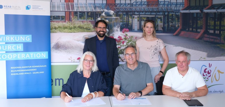 Dr. Katja Wolf, Leiterin der Regionalagentur (vorne links), Landrat Dietmar Seefeldt (Mitte) und Kreisbeigeordneter Werner Schreiner (rechts) unterzeichneten die Vereinbarung im Beisein von Sebastian Müller, kommunaler Berater, und Projektleiterin Monika Kukyte. Foto: KV SÜW 3 Personen sitzen, 2 Personen stehen vor Bannern der Regionalagentur und des Landkreises.