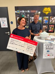Eine Frau hält lächelnd einen großen Spendenscheck über 3062,50 in den Händen. Hinter ihr steht ein ebenfalls lächelnder Mann. Daneben befindet sich eine Spendenbox; im Hintergrund ist eine Präsentationswand von Kissel SBK zu sehen. 