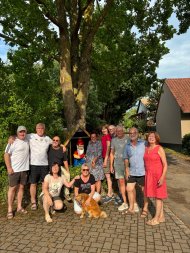 Der Gartenzwerg mit Namen Klaus-Bärbel steht am „Großwiesen Plätzel“ in Klingenmünster für eine gute Nachbarschaft. Foto: Ursel Schoone/frei Eine Gruppe von elf Personen posiert im Freien unter einem großen Baum für ein gemeinsames Foto. Im Hintergrund steht eine Gartenzwergfigur von einem kleinen Holzhäuschen.