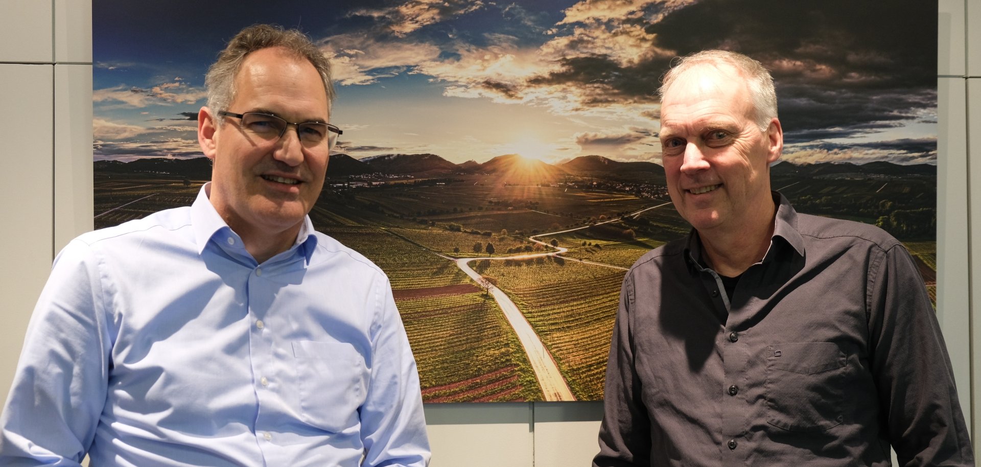 Zwei Männer stehen nebeneinander im Kreishaus Südliche Weinstraße vor einem großformatigen Landschaftsfoto mit Weinbergen. Links Landrat Dietmar Seefeldt, rechts Philipp Jähne. Beide blicken in die Kamera und lächeln.