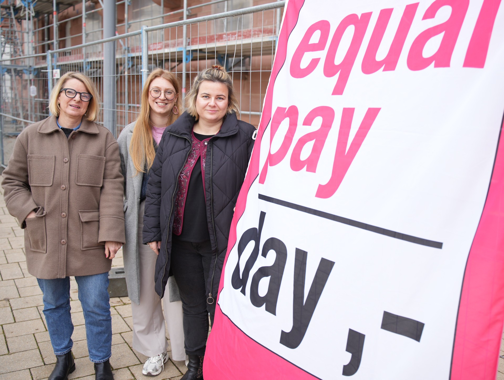 Drei Frauen stehen vor einem Bauzaun und neben einem großen Banner mit der Aufschrift „Equal Pay Day“ und blicken in die Kamera.
