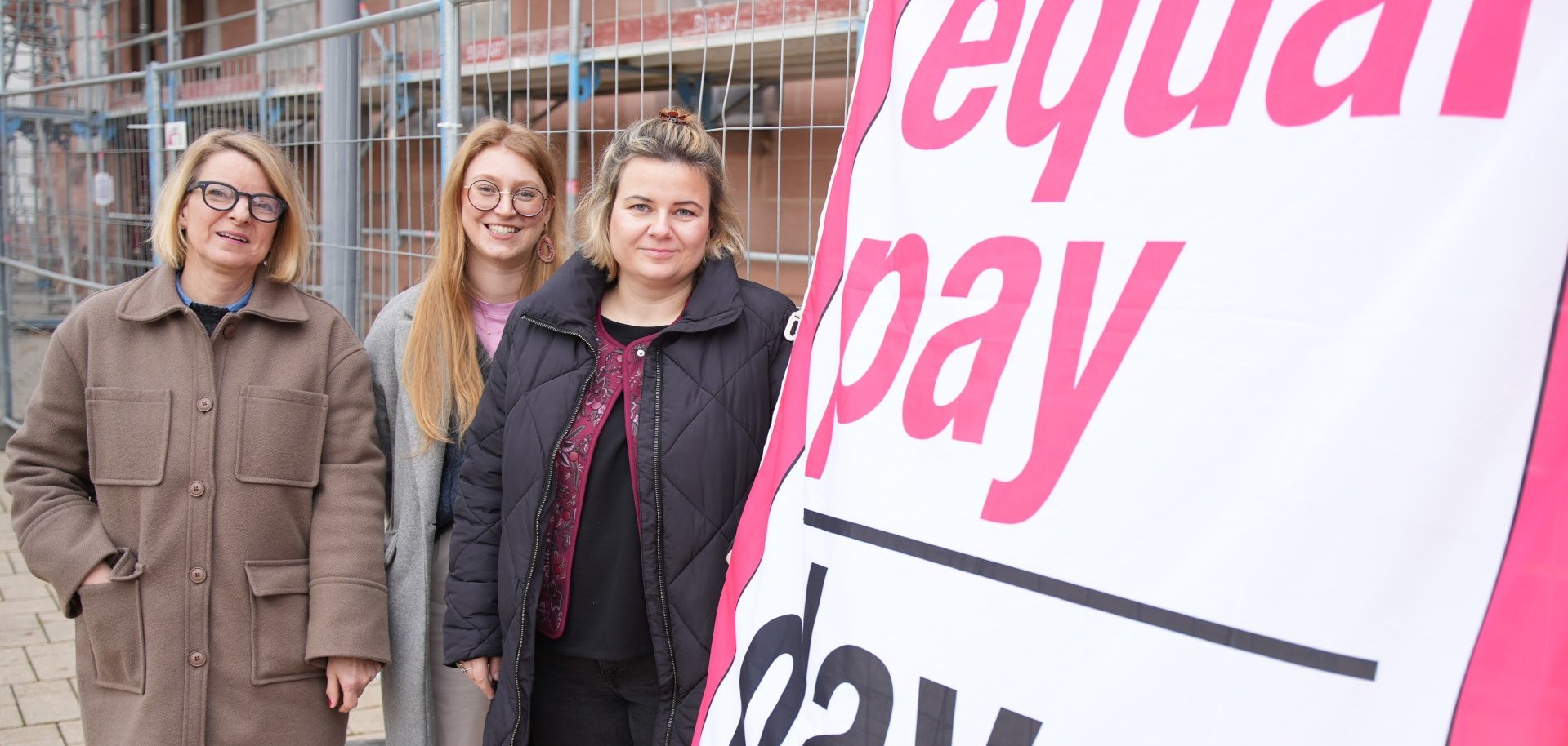 Drei Frauen stehen vor einem Bauzaun und neben einem großen Banner mit der Aufschrift „Equal Pay Day“ und blicken in die Kamera.