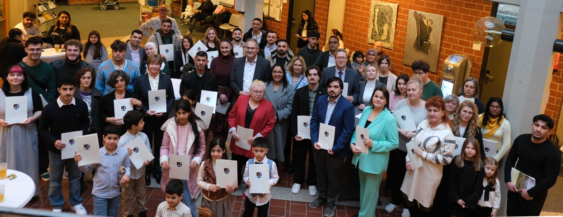 Zahlreiche frisch eingebürgerte deutsche Staatsbürgerinnen und Staatsbürger stehen zusammen im Foyer des Kreishauses der Südlichen Weinstraße, blicken in die Kamera und halten ihre Urkunde in Händen.