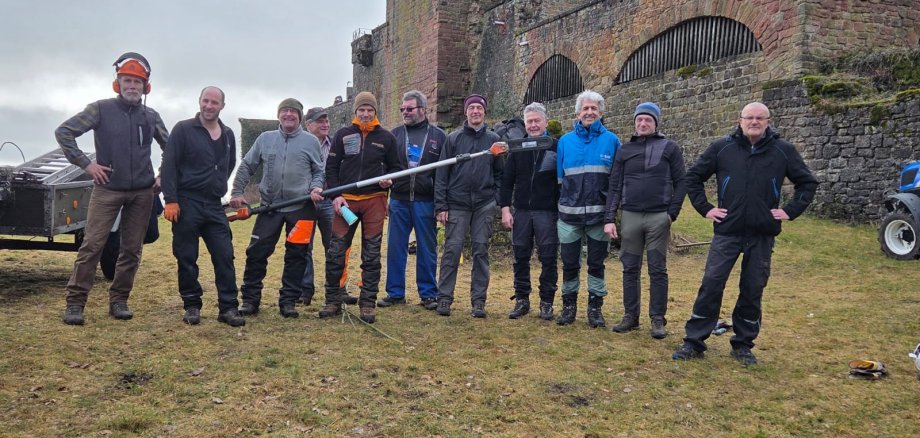Mehrere Männer stehen bei einem Arbeitseinsatz vor der Madenburg und halten gemeinsam eine motorisierte Heckenschere. Im Hintergrund sind die historischen Mauern der Burgruine zu sehen.