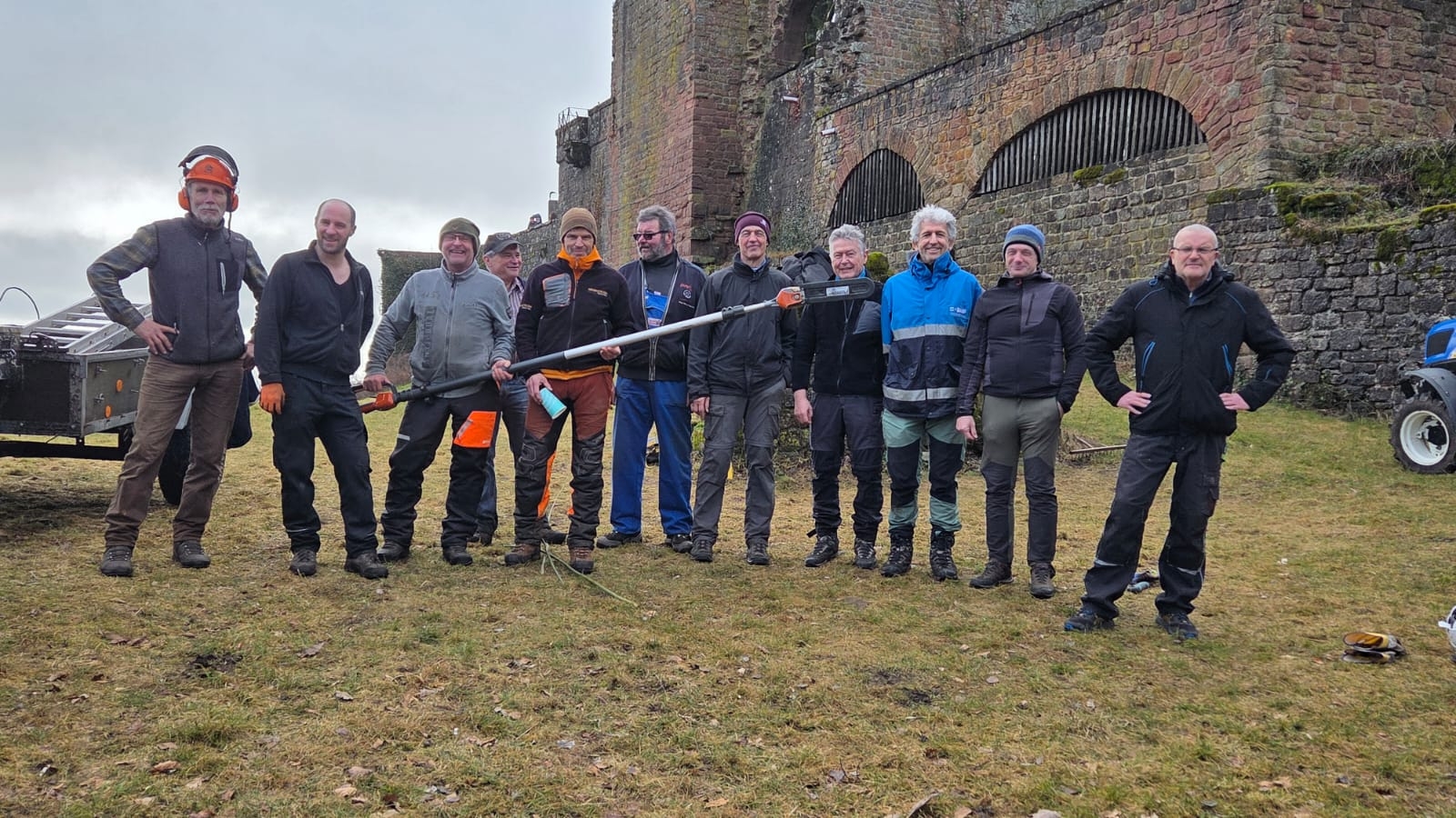 Mehrere Männer stehen bei einem Arbeitseinsatz vor der Madenburg und halten gemeinsam eine motorisierte Heckenschere. Im Hintergrund sind die historischen Mauern der Burgruine zu sehen.