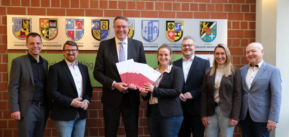 Sieben Personen vor einer Wand mit Wappen der Verbandsgemeinden des Kreises und dem Wappen des Landkreises SÜW. 