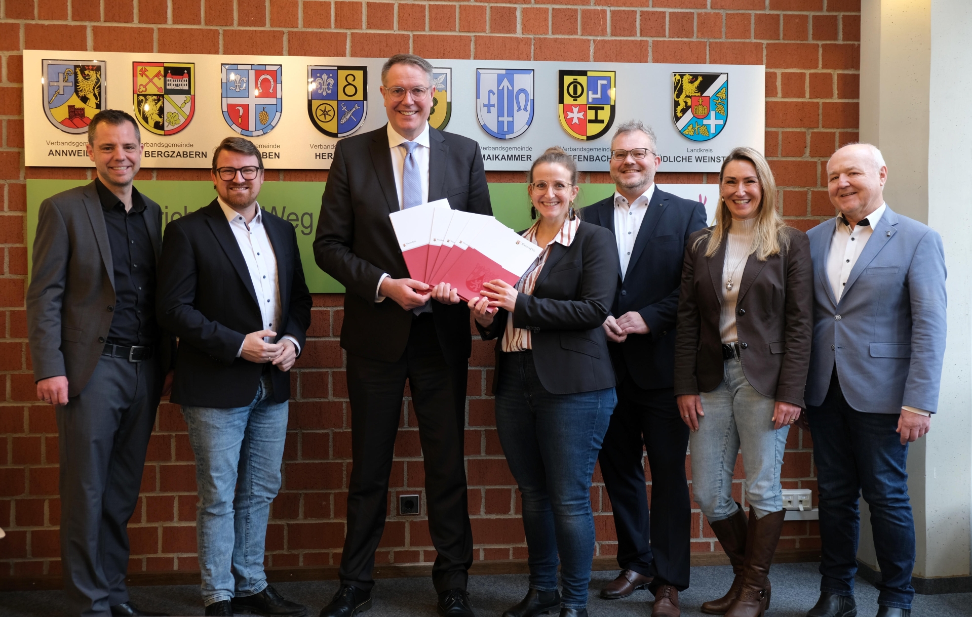 Sieben Personen vor einer Wand mit Wappen der Verbandsgemeinden des Kreises und dem Wappen des Landkreises SÜW. 