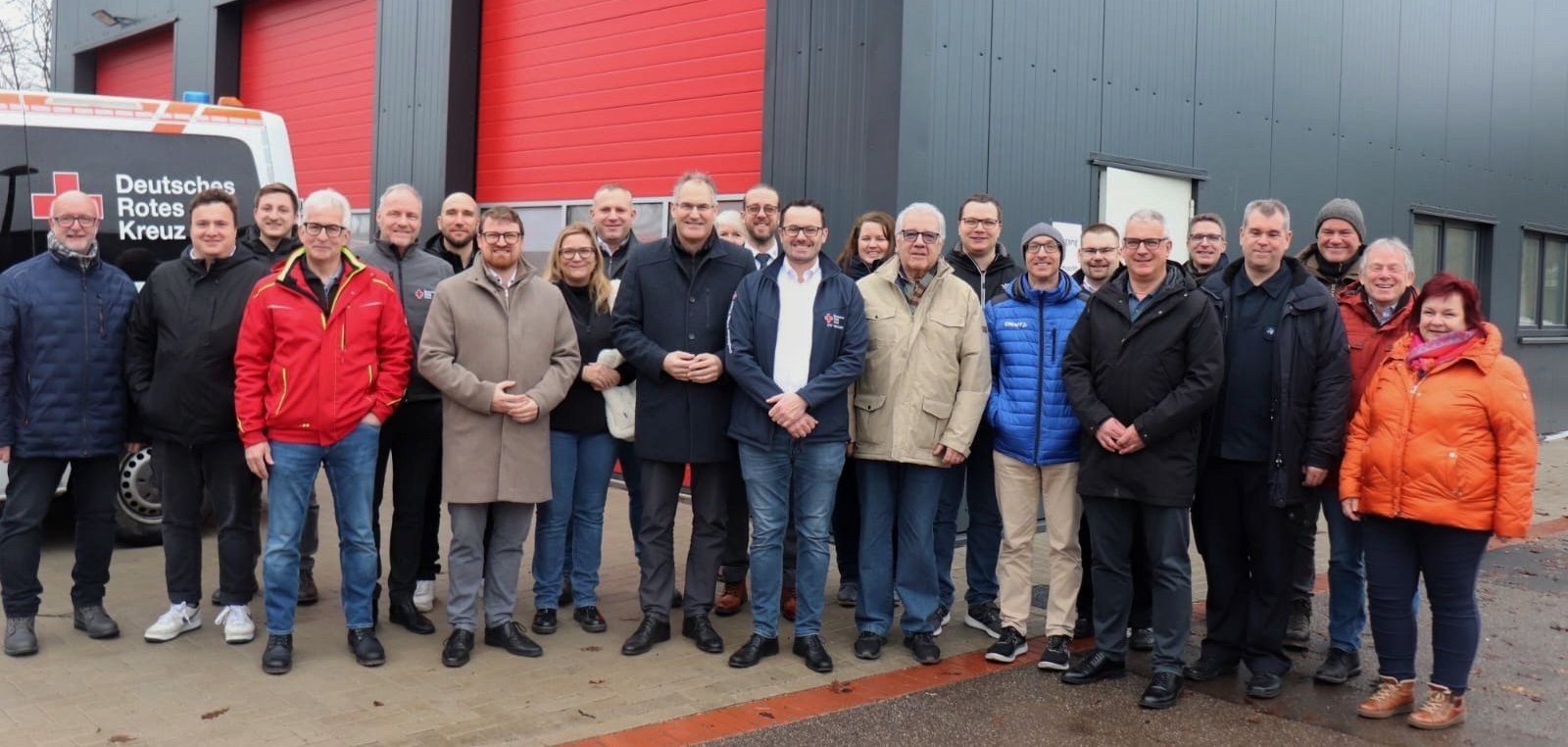 Gruppenbild: Vertreterinnen und Vertreter der örtlichen Politik, Verwaltung und des DRK stehen gemeinsam vor der neuen Halle, von der im Hintergrund unter anderem drei große rote Tore zu sehen sind.