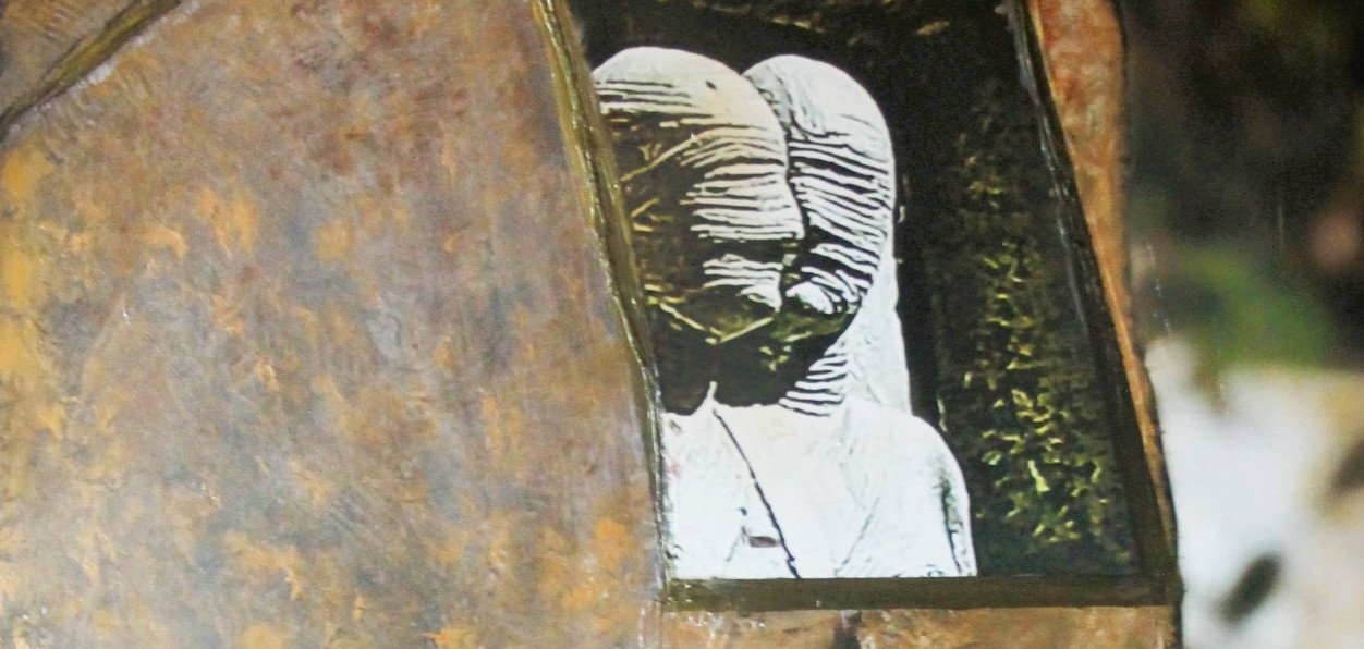 Detailansicht einer Skulptur aus rostfarbenem Metall. In einer Öffnung im Oberkörper ist eine kleine, helle Figur oder Darstellung eines Menschenpaares zu sehen, die einen starken Kontrast zur dunklen, rauen Oberfläche bildet.