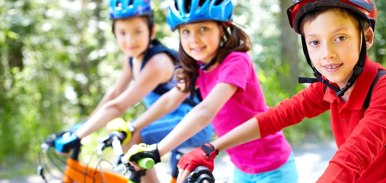 Drei Kinder fahren mit Fahrrädern auf einem Weg im Grünen. Sie tragen Fahrradhelme und bunte Kleidung und blicken lächelnd in die Kamera.