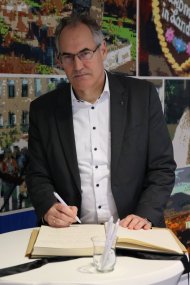 Landrat Dietmar Seefeldt, im dunklen Anzug und weißem Hemd, trägt sich sitzend in das aufgeschlagene Kondolenzbuch ein. Im Hintergrund ist eine großformatige, farbige Bildcollage zu sehen.