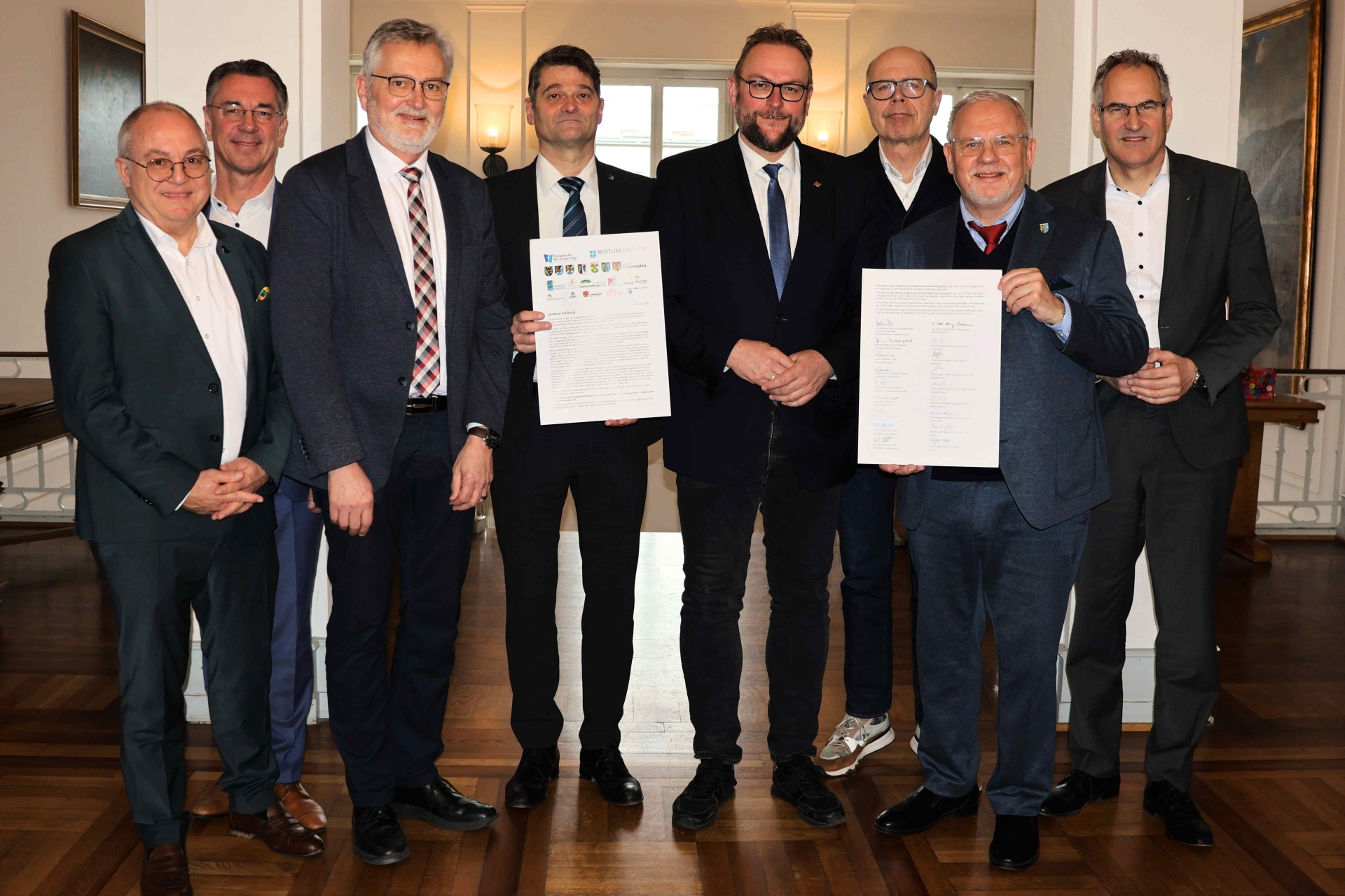 Mehrere Männer in Anzügen stehen in einem repräsentativen Raum nebeneinander. Zwei von ihnen halten jeweils ein Dokument mit Unterschriften in die Kamera.