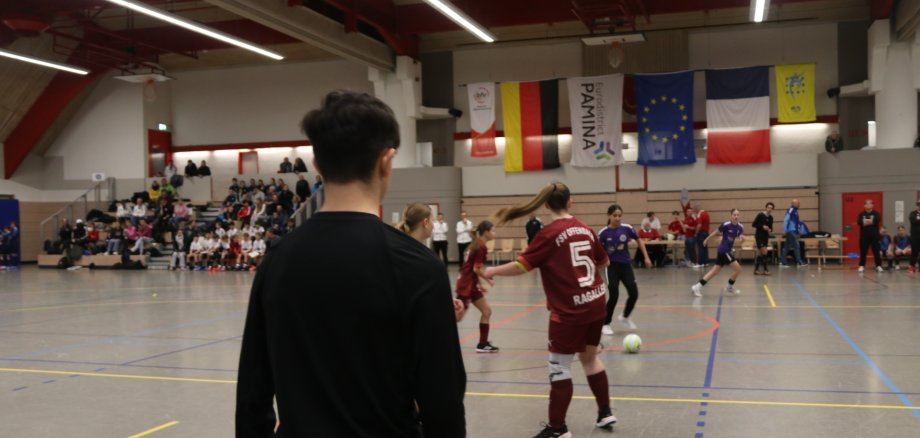 Beim PAMINA-Cup 2026 traten C-Juniorinnen und D-Junioren aus Deutschland und Frankreich gegeneinander an. Foto: Badischer Fußballverband e.V. Eine Spielsituation beim PAMINA-Cup in einer Sporthalle: Jugendliche Fußballspielerinnen und -spieler stehen während eines Spiels auf dem Hallenfeld, im Hintergrund verfolgen Zuschauerinnen und Zuschauer das Turnier von der Tribüne. Über dem Spielfeld hängen Fahnen verschiedener Länder und Regionen.