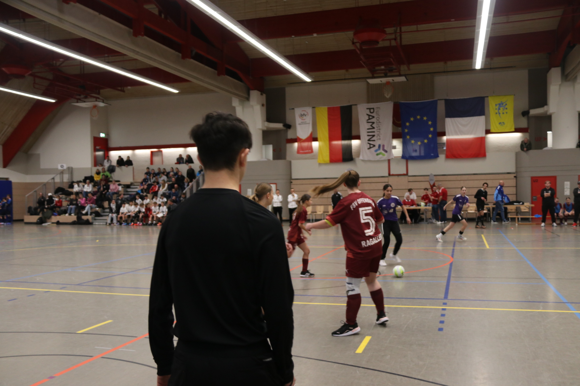 Eine Spielsituation beim PAMINA-Cup in einer Sporthalle: Jugendliche Fußballspielerinnen und -spieler stehen während eines Spiels auf dem Hallenfeld, im Hintergrund verfolgen Zuschauerinnen und Zuschauer das Turnier von der Tribüne. Über dem Spielfeld hängen Fahnen verschiedener Länder und Regionen.
