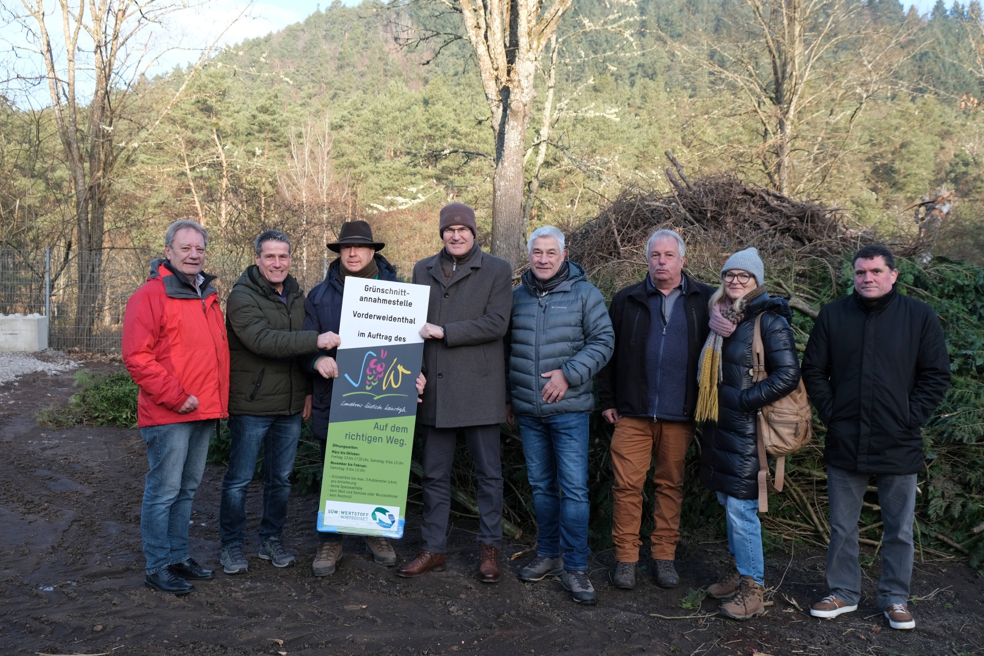 Acht Personen im Freien vor einem Berg Grünschnitt. Drei von ihnen halten ein Schild mit der Aufschrift "Grünschnitt-Annahmestelle Vorderweidenthal."