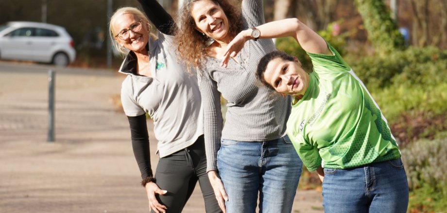Drei Frauen stehen im Freien auf einem gepflasterten Weg und machen gemeinsam eine seitliche Dehnübung. Sie neigen ihre Oberkörper zur Seite und strecken jeweils einen Arm nach oben. Die Frauen lächeln in die Kamera, im Hintergrund sind Bäume, Straßenlampen und geparkte Autos zu sehen.