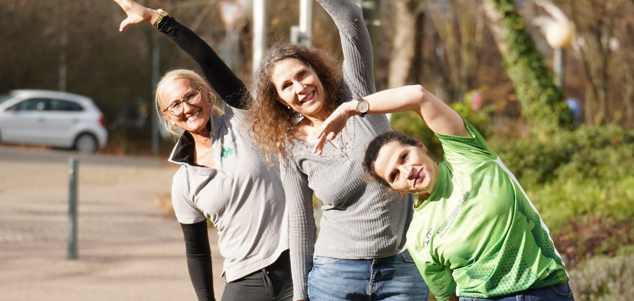 Drei Frauen stehen im Freien auf einem gepflasterten Weg und machen gemeinsam eine seitliche Dehnübung. Sie neigen ihre Oberkörper zur Seite und strecken jeweils einen Arm nach oben. Die Frauen lächeln in die Kamera, im Hintergrund sind Bäume, Straßenlampen und geparkte Autos zu sehen.