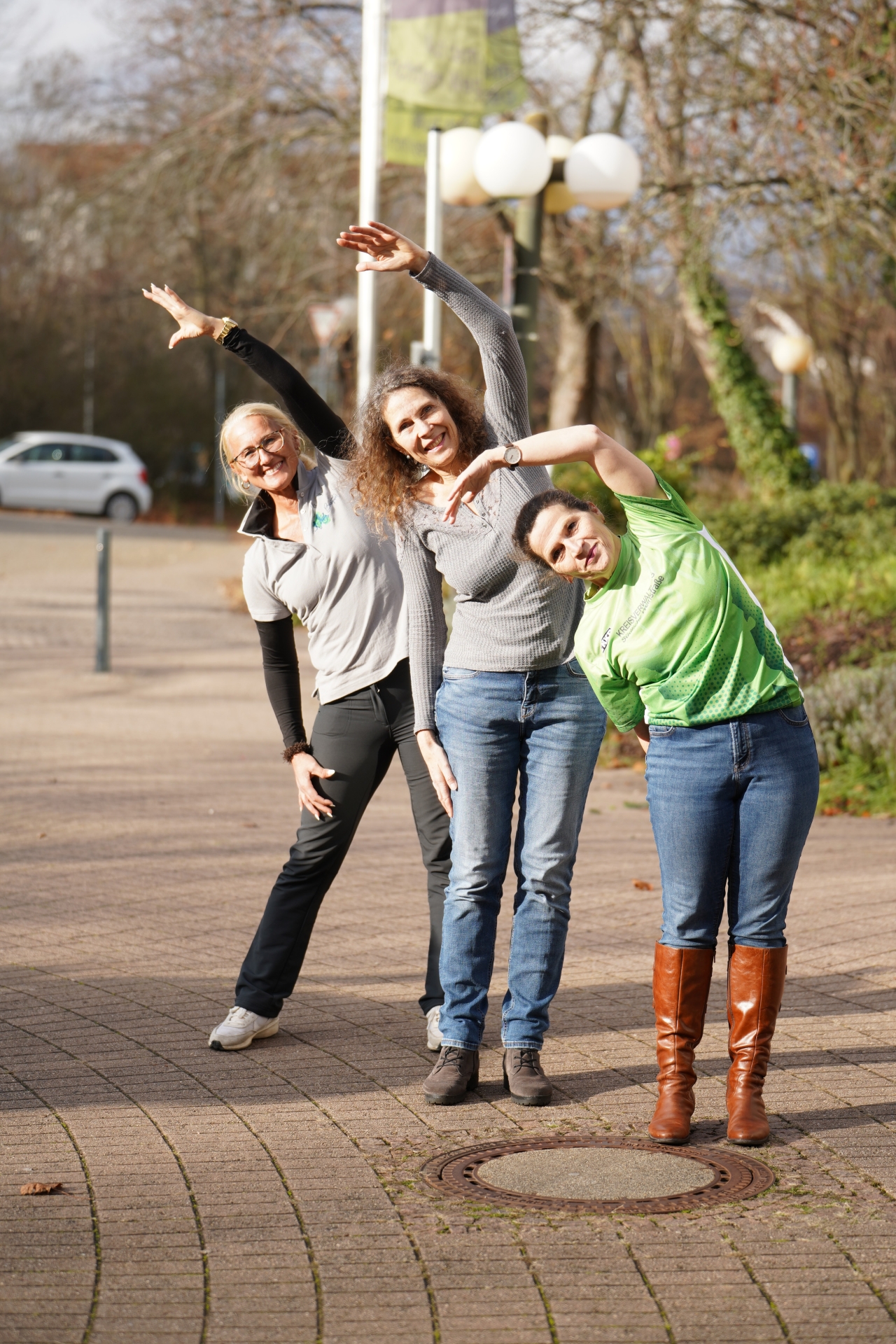 Drei Frauen stehen im Freien auf einem gepflasterten Weg und machen gemeinsam eine seitliche Dehnübung. Sie neigen ihre Oberkörper zur Seite und strecken jeweils einen Arm nach oben. Die Frauen lächeln in die Kamera, im Hintergrund sind Bäume, Straßenlampen und geparkte Autos zu sehen.