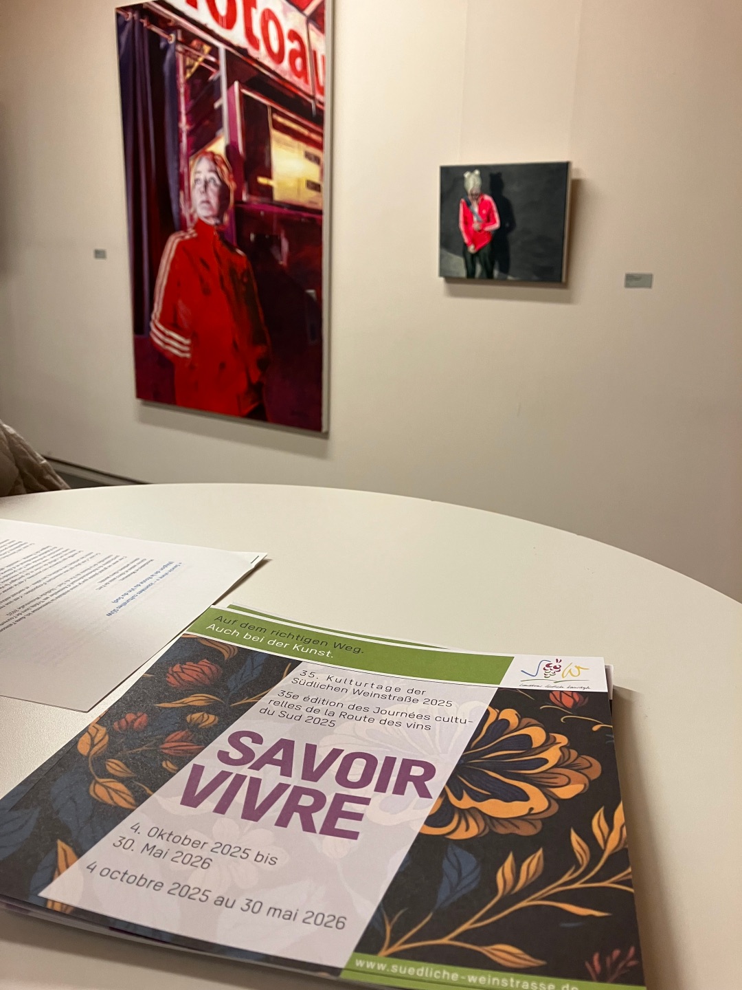 Auf einem runden Tisch liegt ein aufgeschlagenes Programmheft mit der Aufschrift „Savoir vivre – Kulturtage der Südlichen Weinstraße 2025“. Im Hintergrund sind an einer hellen Galeriewand zwei Kunstwerke zu sehen: links ein großformatiges Porträt einer Person in roter Jacke, rechts ein kleineres Bild mit einer stehenden Figur vor dunklem Hintergrund.