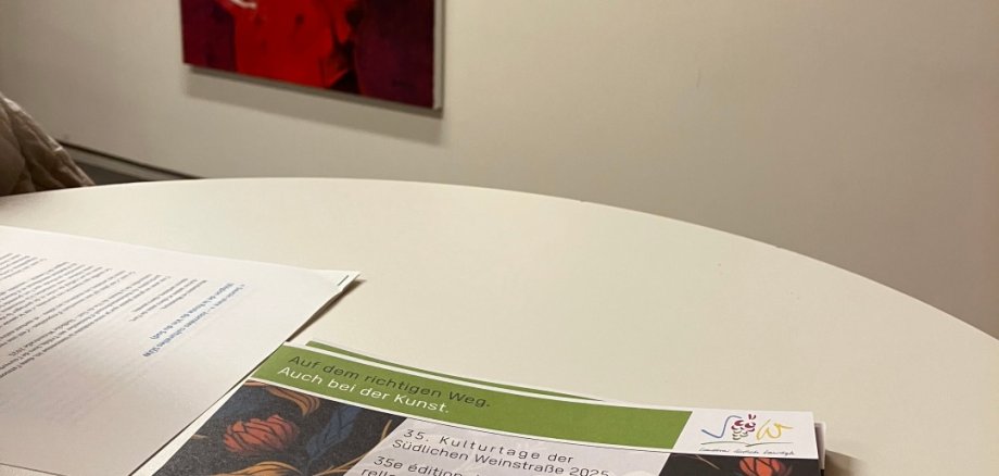 Auf einem runden Tisch liegt ein aufgeschlagenes Programmheft mit der Aufschrift „Savoir vivre – Kulturtage der Südlichen Weinstraße 2025“. Im Hintergrund sind an einer hellen Galeriewand zwei Kunstwerke zu sehen: links ein großformatiges Porträt einer Person in roter Jacke, rechts ein kleineres Bild mit einer stehenden Figur vor dunklem Hintergrund.