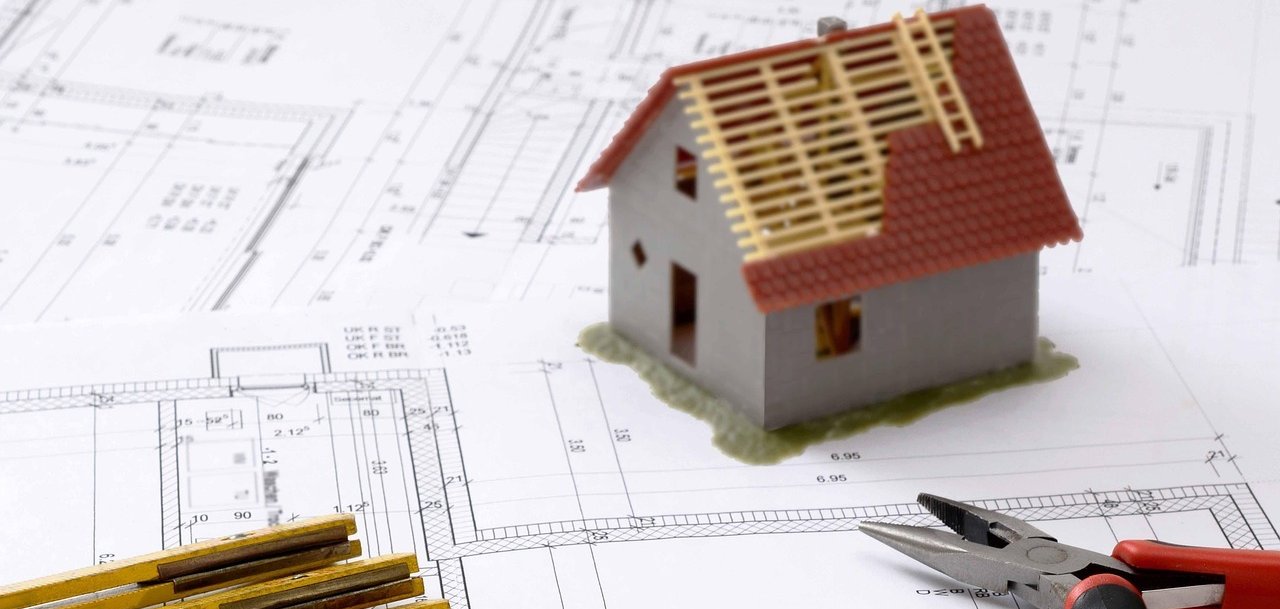 Modell eines Einfamilienhauses steht auf Bauplänen. Daneben liegen Zollstock, Bleistift und Zange als Symbole für Planung und Bauvorhaben.