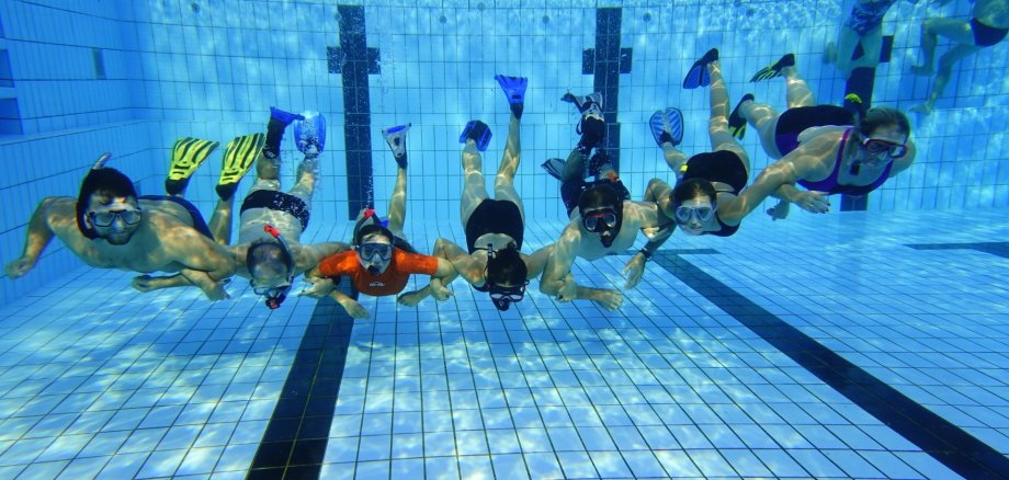 Mehrere Taucherinnen und Taucher halten sich unter Wasser an den Händen und schauen in die Kamera. Alle haben Taucherbrillen auf.