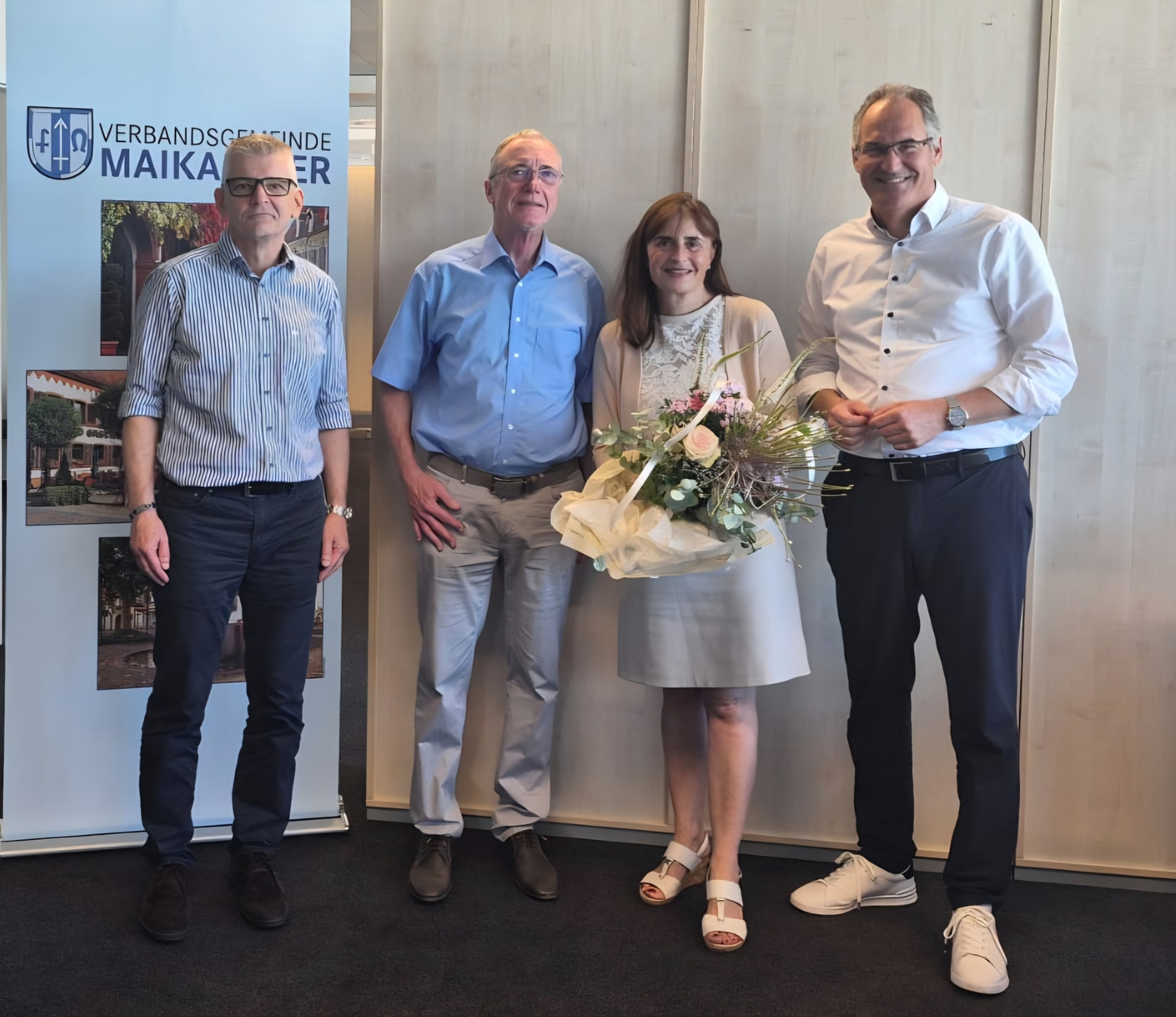 Vier Personen stehen bei einer Feierstunde nebeneinander. Eine Frau hält einen großen Blumenstrauß und wird von drei Männern flaniert. Im Hintergrund ist ein Roll-up der Verbandsgemeinde Maikammer zu sehen. Das Bild zeigt eine Ehrung oder Verabschiedung im offiziellen Rahmen.