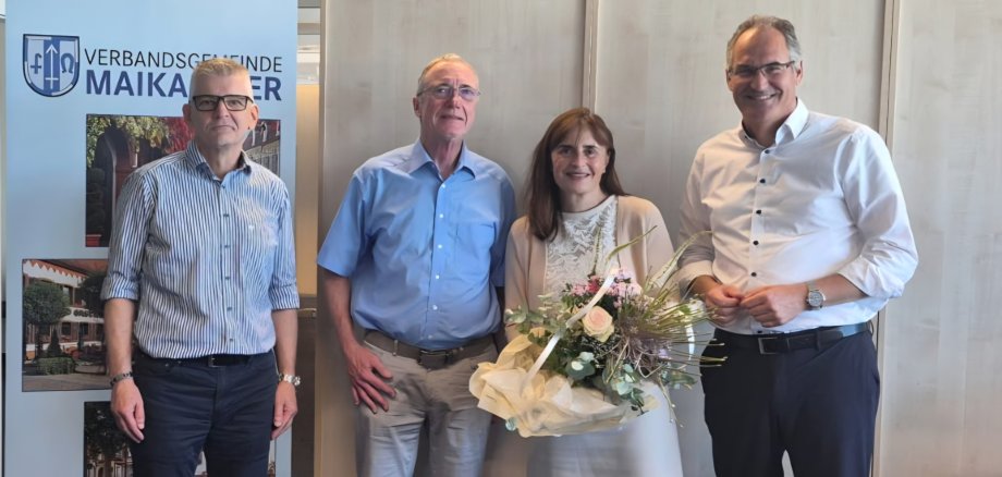 Bürgermeisterin Gabriele Flach (mit Blumen) im Kreis ihrer Gratulanten: Büroleiter Andreas Reuter, Erster Beigeordneter Dr. Reinhold Stadler und Landrat Dietmar Seefeldt (von links nach rechts). Foto: VG Maikammer Vier Personen stehen bei einer Feierstunde nebeneinander. Eine Frau hält einen großen Blumenstrauß und wird von drei Männern flaniert. Im Hintergrund ist ein Roll-up der Verbandsgemeinde Maikammer zu sehen. Das Bild zeigt eine Ehrung oder Verabschiedung im offiziellen Rahmen.