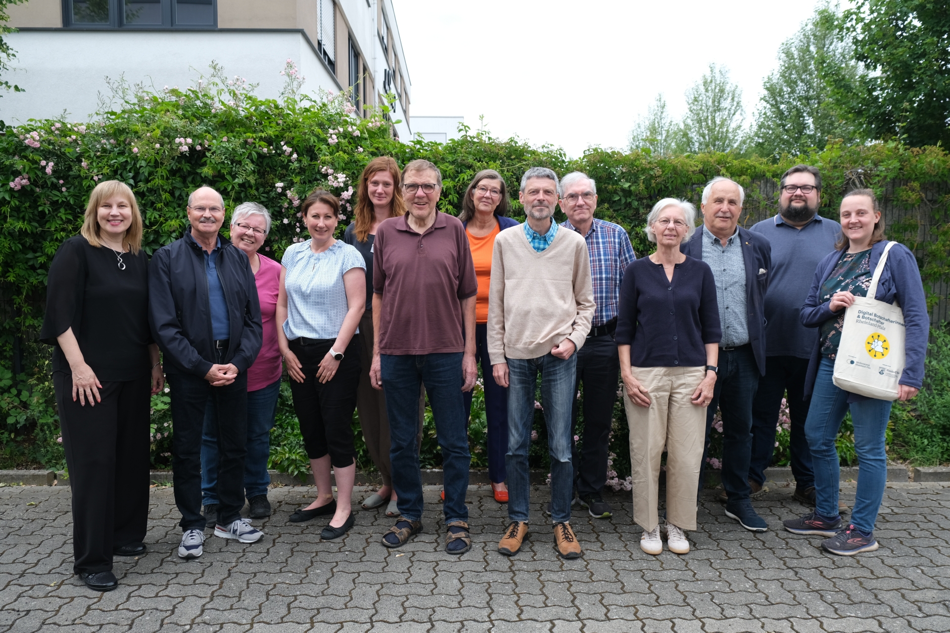Dreizehn Personen stehen für ein Gruppenfoto im Freien vor einer blühenden Hecke.