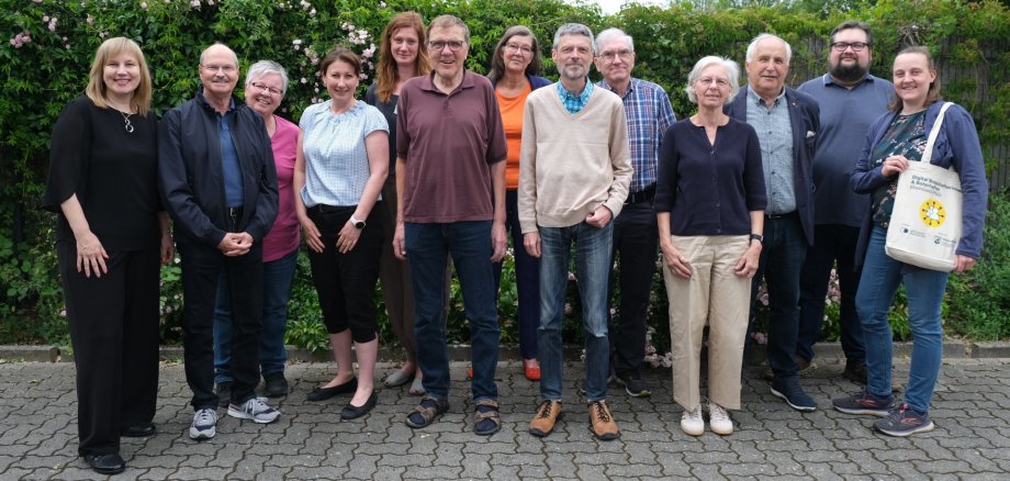 Dreizehn Personen stehen für ein Gruppenfoto im Freien vor einer blühenden Hecke.