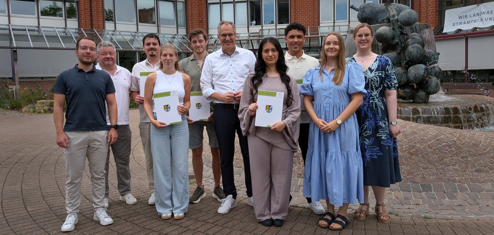 Gruppenfoto vor dem Kreishaus der Kreisverwaltung Südliche Weinstraße: Mehrere Nachwuchskräfte halten ihre Ernennungs- beziehungsweise Ausbildungsunterlagen in den Händen und stehen gemeinsam mit Landrat Dietmar Seefeldt sowie weiteren Mitarbeitenden für ein Foto zusammen.