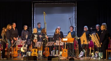 Das Zupforchester der Kreismusikschule SÜW ist im August wieder zu hören, hier eine Aufnahme aus dem Jahr 2023. Foto: KV SÜW Mehrere Musikerinnen und Musiker stehen auf einer Bühne mit ihren Instrumenten. zu sehen sind unter anderem Veeh-Harfen, Gitarren und weitere Instrumente sowie Notenständer und Mikrofone.