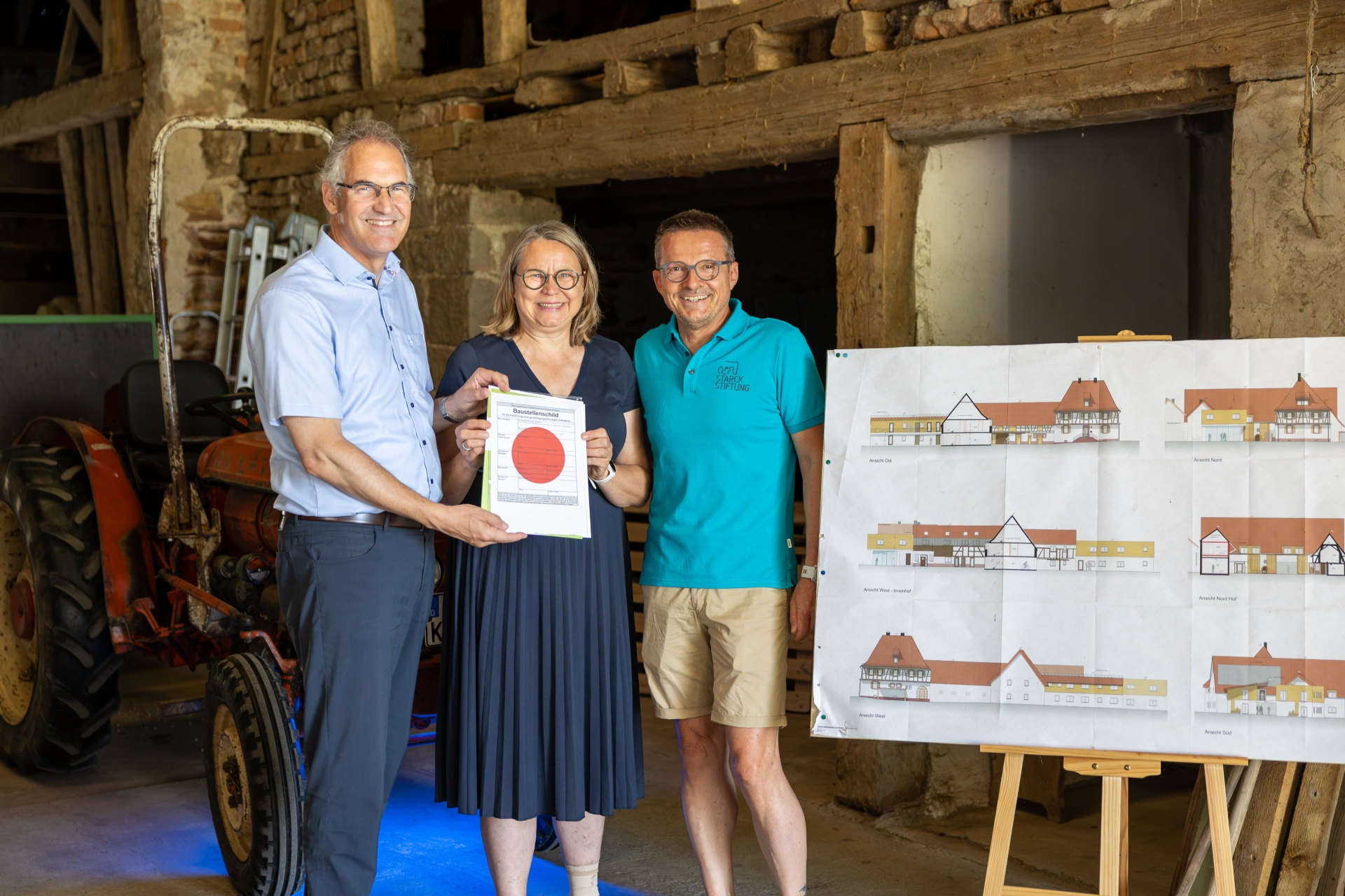 Landrat Dietmar Seefeldt, Astrid und Georg Starck stehen in einer Scheune neben einer Staffelei, auf der eine Abbildung des geplanten Bauvorhabens steht.