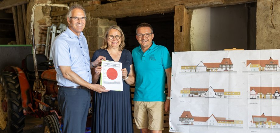 Landrat Dietmar Seefeldt hat Astrid und Georg Starck die Baugenehmigung für den Pflegebauernhof „Hofgut Böhlwiesen“ persönlich überreicht. Rechts im Bild sind die Pläne für das Bauvorhaben zu sehen. Foto: Starck Stiftung Landrat Dietmar Seefeldt, Astrid und Georg Starck stehen in einer Scheune neben einer Staffelei, auf der eine Abbildung des geplanten Bauvorhabens steht.