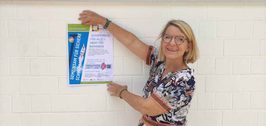 Auch im Trifelsbad Annweiler hat die SÜW-Gleichstellungsbeauftragte Isabelle Stähle Plakate zum Thema Sicherheit in Schwimmbädern angebracht. Foto: Trifelsbad Annweiler Die SÜW-Gleichstellungsbeauftragte Isabelle Stähle hat an einer weißen Wand im Trifelsbad in Annweiler ein Plakat zum Thema Sicherheit in Schwimmbädern angebracht.