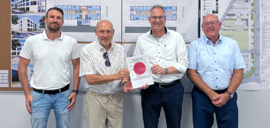 Vier Männer stehen vor einer Pinnwand mit Bauplänen und Architekturvisualisierungen. Zwei von ihnen halten gemeinsam ein Dokument mit einer Baugenehmigung in die Kamera. Die Gruppe lächelt und posiert für das Foto in einem Besprechungsraum.