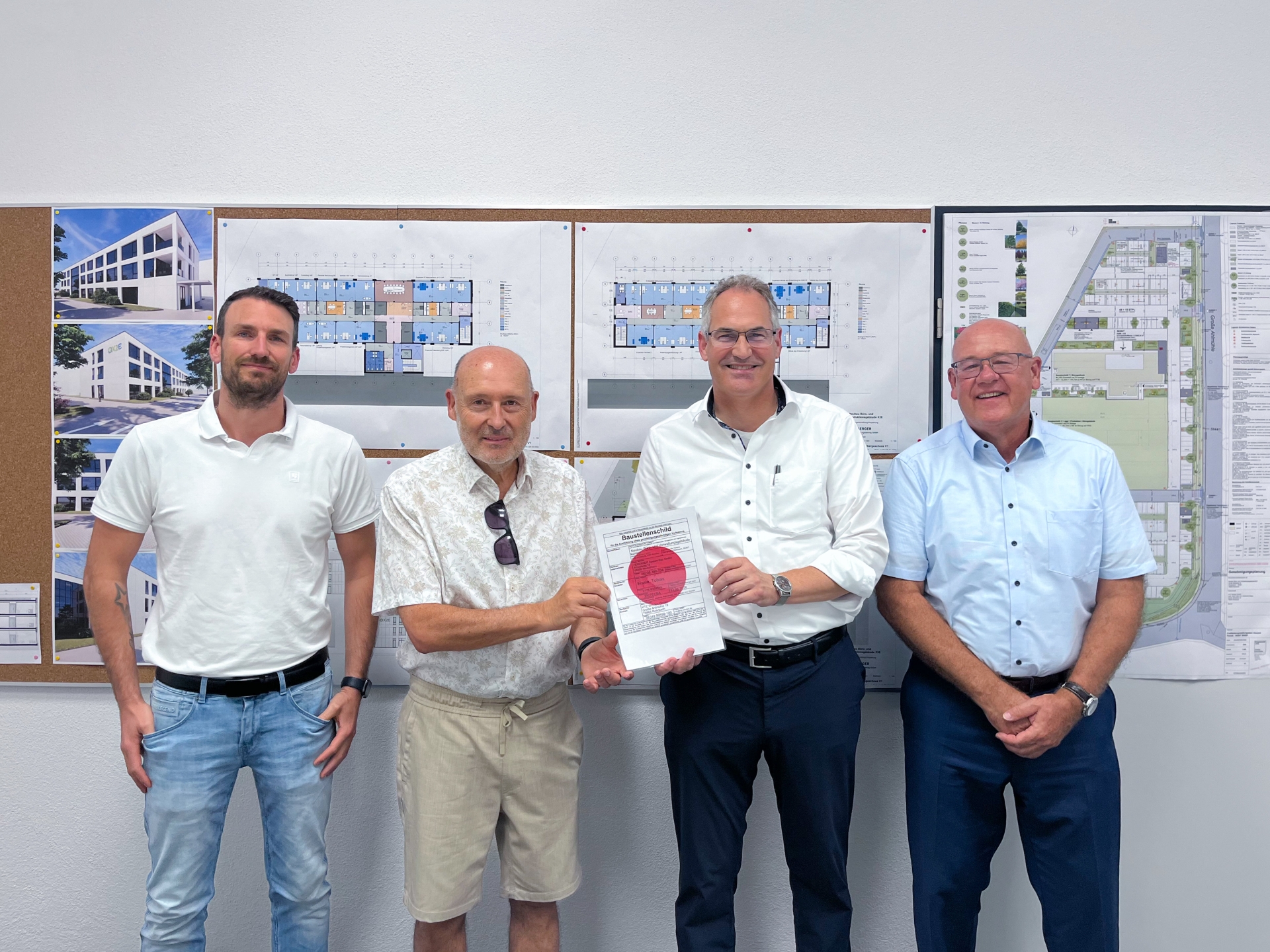 Vier Männer stehen vor einer Pinnwand mit Bauplänen und Architekturvisualisierungen. Zwei von ihnen halten gemeinsam ein Dokument mit einer Baugenehmigung in die Kamera. Die Gruppe lächelt und posiert für das Foto in einem Besprechungsraum.
