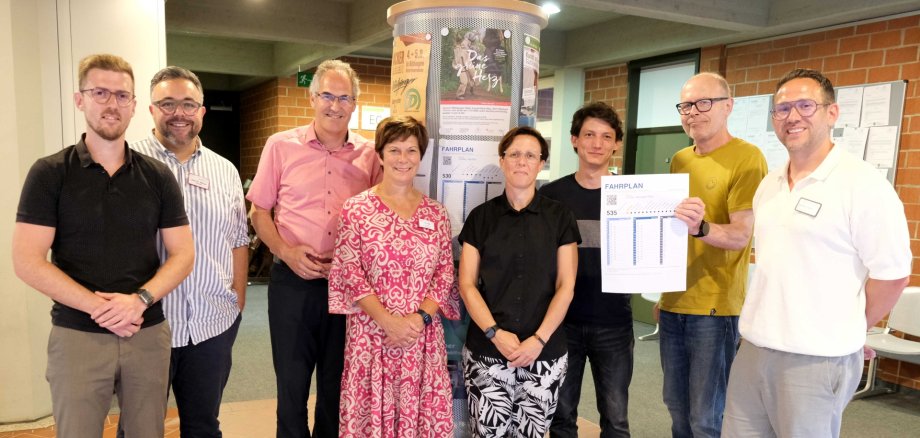 Tauschten sich zum Thema ÖPNV-Verbesserungen für Pflege-Azubis aus: Benjamin Klar, ÖPNV-Referent Landkreis SÜW, Dr. Marvin Milius (Leiter Pflegeschule Klinikum Landau-SÜW), SÜW-Landrat Dietmar Seefeldt, Monika Vogler (Schulleitung Nardini Pflegeschule), Kathrin Flory, Bürgermeisterin der Verbandsgemeinde Bad Bergzabern, Stefan Grewenig, ÖPNV-Referent der Stadt Landau, Landaus Oberbürgermeister Dominik Geißler, und Sebastian Löhlein (Leiter Bildungszentrum Pfalzklinikum). Auf dem Foto nicht zu sehen ist Landrat Martin Brandl (GER), der ebenfalls teilnahm, allerdings kurz vor der Aufnahme zu einem Anschlusstermin musste. Foto: KV SÜW Acht Personen stehen im Foyer der Kreisverwaltung vor einer Litfaßsäule, Dominik Geißler hält einen Fahrplan der Landauer ÖPNV-Linie 535 hoch.