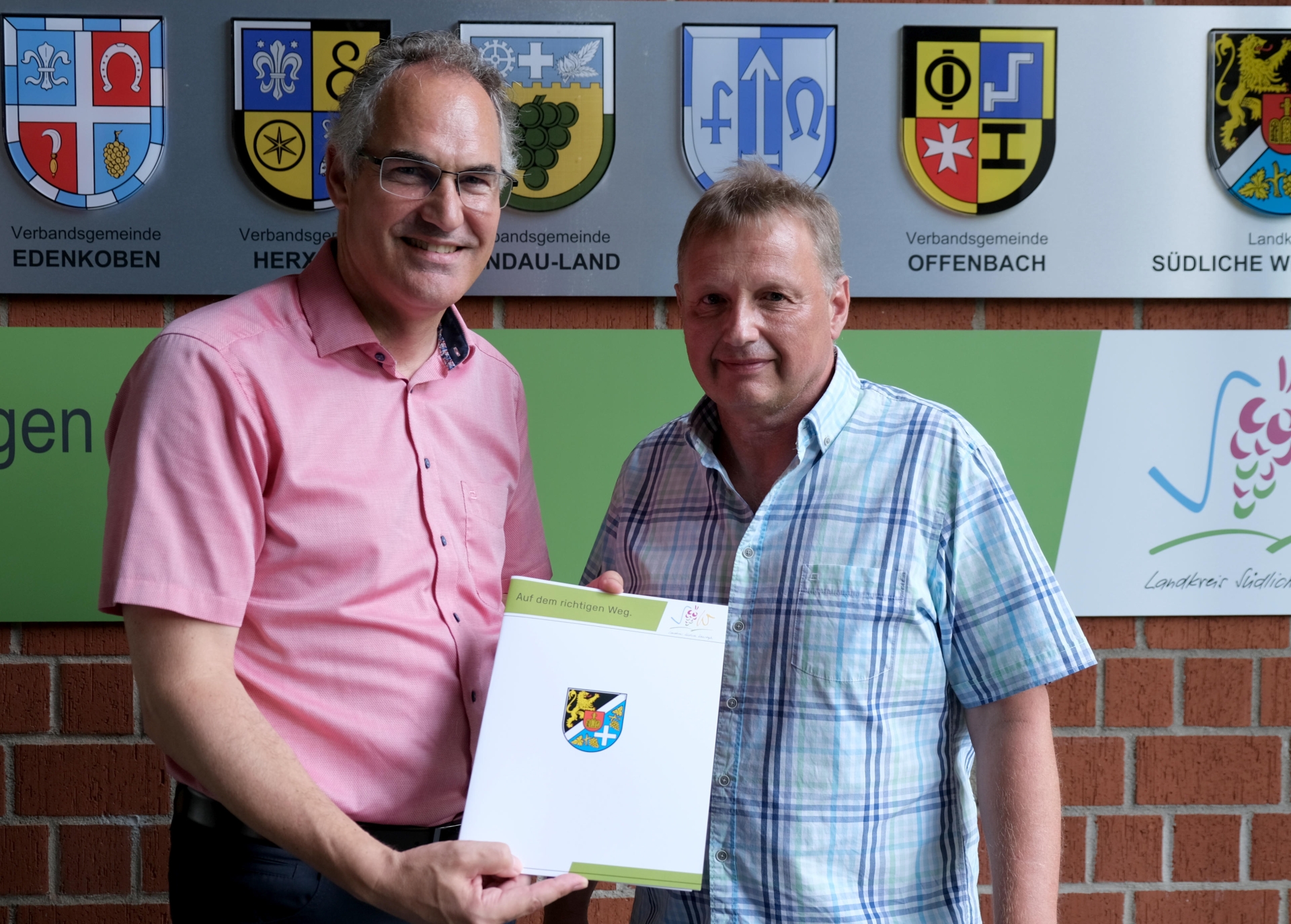 Landrat Dietmar Seefeldt und Ralf Stolz im Porträt. Landrat Seefeldt hält eine Mappe mit dem Wappen des Landkreises SÜW.