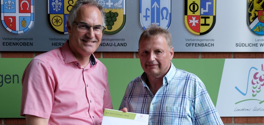 Ralf Stolz (rechts) wurde von Landrat Dietmar Seefeldt in den Ruhestand verabschiedet. Foto: KV SÜW Landrat Dietmar Seefeldt und Ralf Stolz im Porträt. Landrat Seefeldt hält eine Mappe mit dem Wappen des Landkreises SÜW.