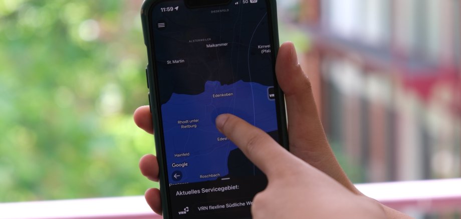 In Rhodt unter Rietburg und Edenkoben gibt es neue virtuelle Haltestellen der VRNflexline. Foto: KV SÜW Nahaufnahme von Händen mit einem Smartphone. Jemand zeigt auf den Bildschirm, auf dem eine Karte der Verbandsgemeinde Edenkoben in der VRNflexline-App zu sehen ist, im Hintergrund grüne Blätter und ein Gebäude.