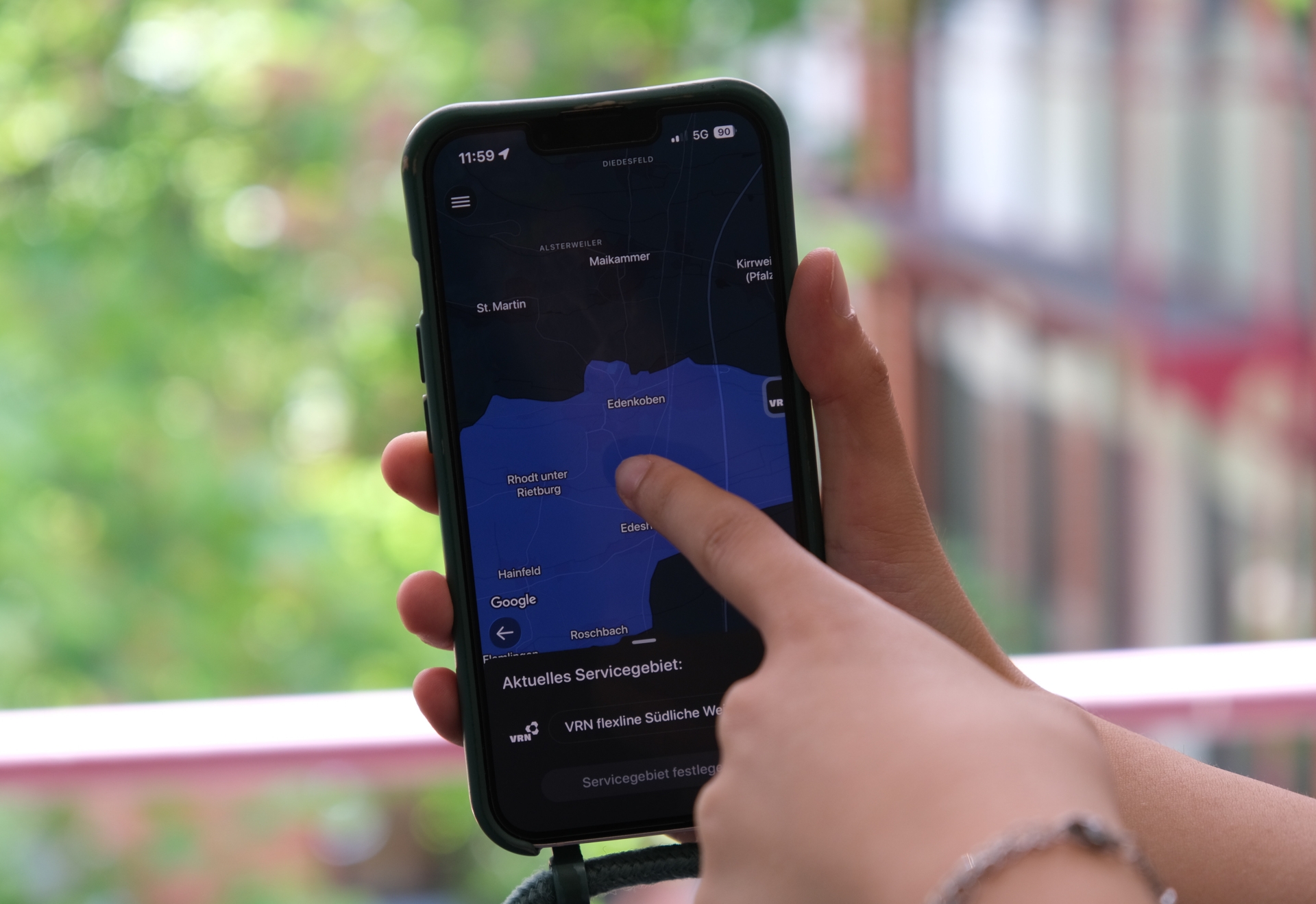 Nahaufnahme von Händen mit einem Smartphone. Jemand zeigt auf den Bildschirm, auf dem eine Karte der Verbandsgemeinde Edenkoben in der VRNflexline-App zu sehen ist, im Hintergrund grüne Blätter und ein Gebäude.