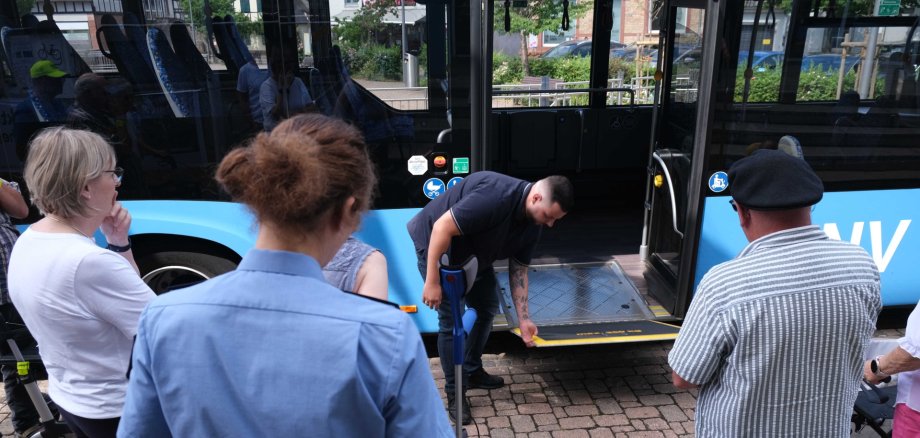 Personen mit Rollator warten, während ein Mann eine Rampe aus dem stehenden Bus ausklappt.