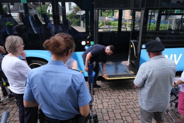 Busfahrer Cevin Klein von der QNV (Mitte) zeigte, wie Ein- und Aussteigen in einen Bus auch mit Rollator gut funktionieren. Foto: KV SÜW Personen mit Rollator warten, während ein Mann eine Rampe aus dem stehenden Bus ausklappt.