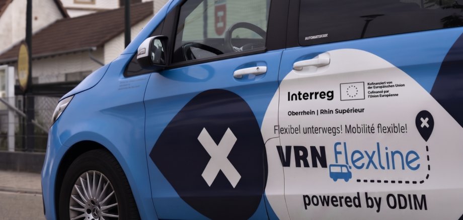So könnten die Kleinbusse auf Abruf aussehen, die grenzübergreifend unterwegs sein werden. Fotomontage mit KI: KV SÜW Ein blauer Kleinbus mit Interreg-Logo, im Hintergrund Häuser mit Nestern mit Weißstörchen.
