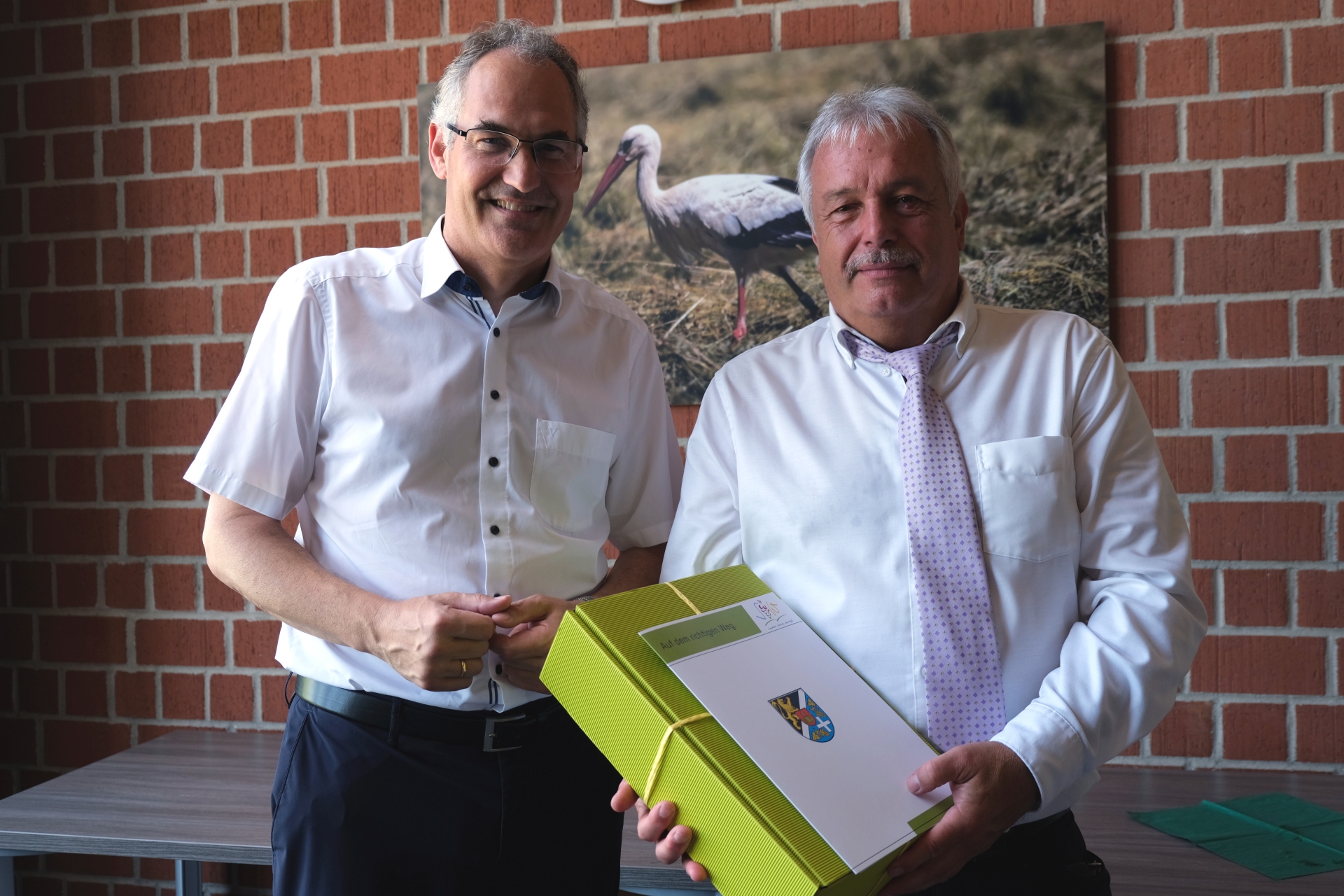 Landrat Dietmar Seefeldt und Rolf Mäckel nebeneinander im Porträt. Rolf Mäckel hält einen grünen Präsentkarton sowie eine Mappe mit Logo und Wappen des Landkreises SÜW in Händen.